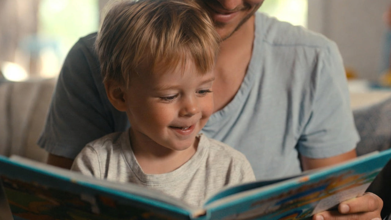 Man and blonde child reading a book together in a cozy living room.
